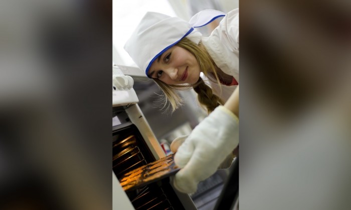 Zdjęcie prezentujące kierunek technik żywienia i usług gastronomicznych. Zdjęcie prezentuje uśmiechającą się, młodą dziewczynę wkładającą blachę z ciastem do piekarnika.