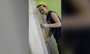 Zdjęcie prezentuje kierunek technik budownictwa. Zdjęcie przedstawia uśmiechającego się młodego chłopaka w kasku. Trzyma on poziomicę murarską przy ścianie.
